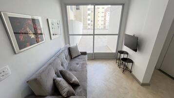 Een moderne woonkamer met een grijze bank, een aan de muur bevestigde televisie en een groot raam met uitzicht op gebouwen.
