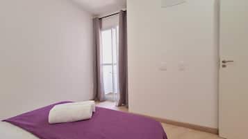 Una habitación con cama, una puerta, un ventanal con cortinas y un interruptor de luz en la pared.