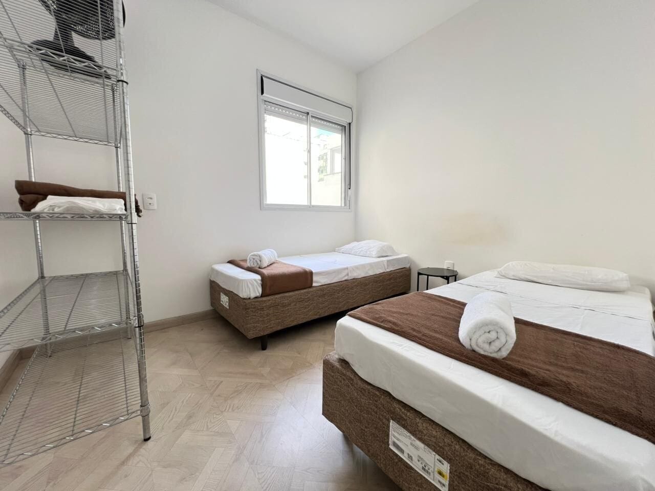 Ein Zimmer mit zwei Betten, einem Metallregal, einem Fenster und einem kleinen Tisch.