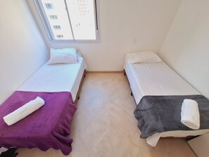 A room with two beds, each with a pillow and a blanket. There is a window with a view of buildings outside.