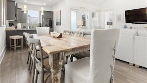 A modern kitchen with a dining area, white furniture, and a large TV. - Frangista Beach: Whispering Palms (Miramar Beach)