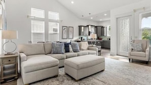 A spacious living room with a large sectional sofa, a small ottoman, and a dining area with a kitchen in the background. - Frangista Beach: Whispering Palms (Miramar Beach)