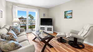 A living room with a sofa, a glass coffee table, a TV on a cabinet, a reclining chair, and a balcony with a view of a pool and buildings. - Tradewinds Condominium #25 (Miramar Beach)