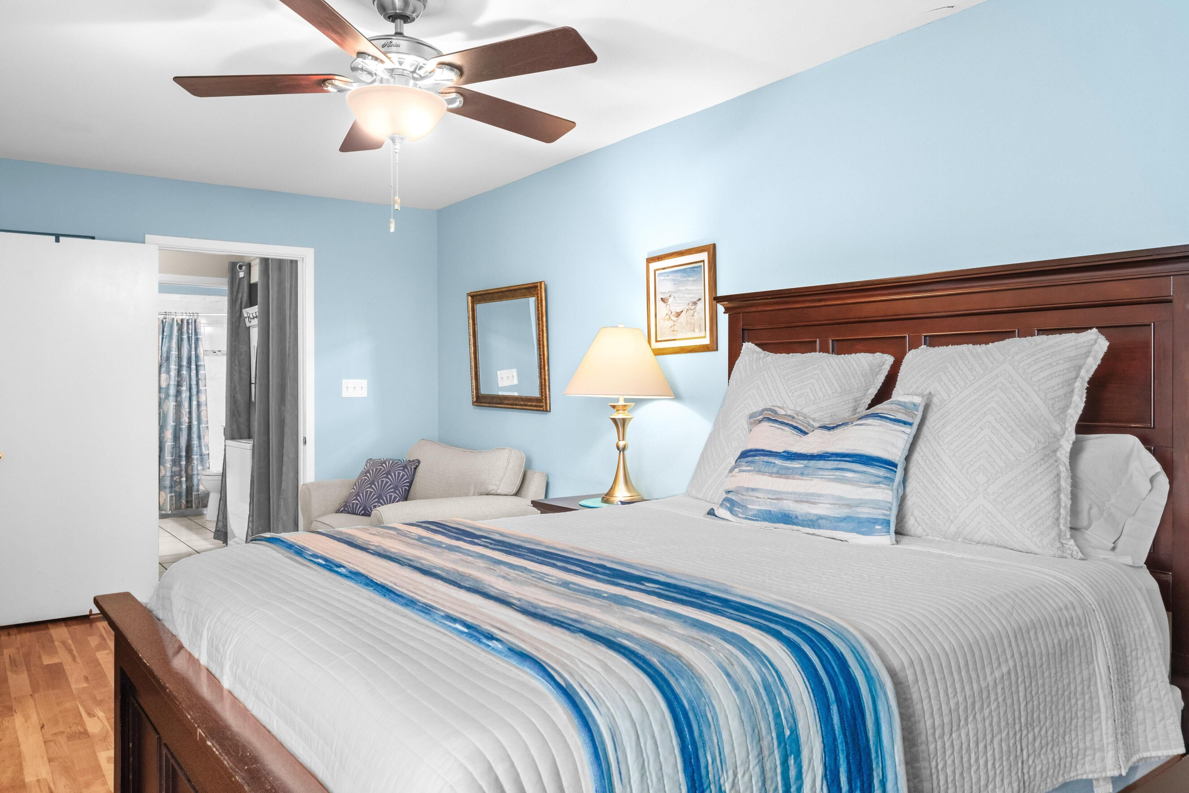 A bedroom with a bed, a ceiling fan, a nightstand with a lamp, and a door leading to a bathroom.