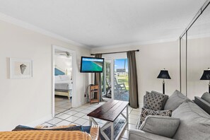 A living room with a sofa, a table, a TV, and a balcony with a view. - Tradewinds Condominium #2 (Miramar Beach)