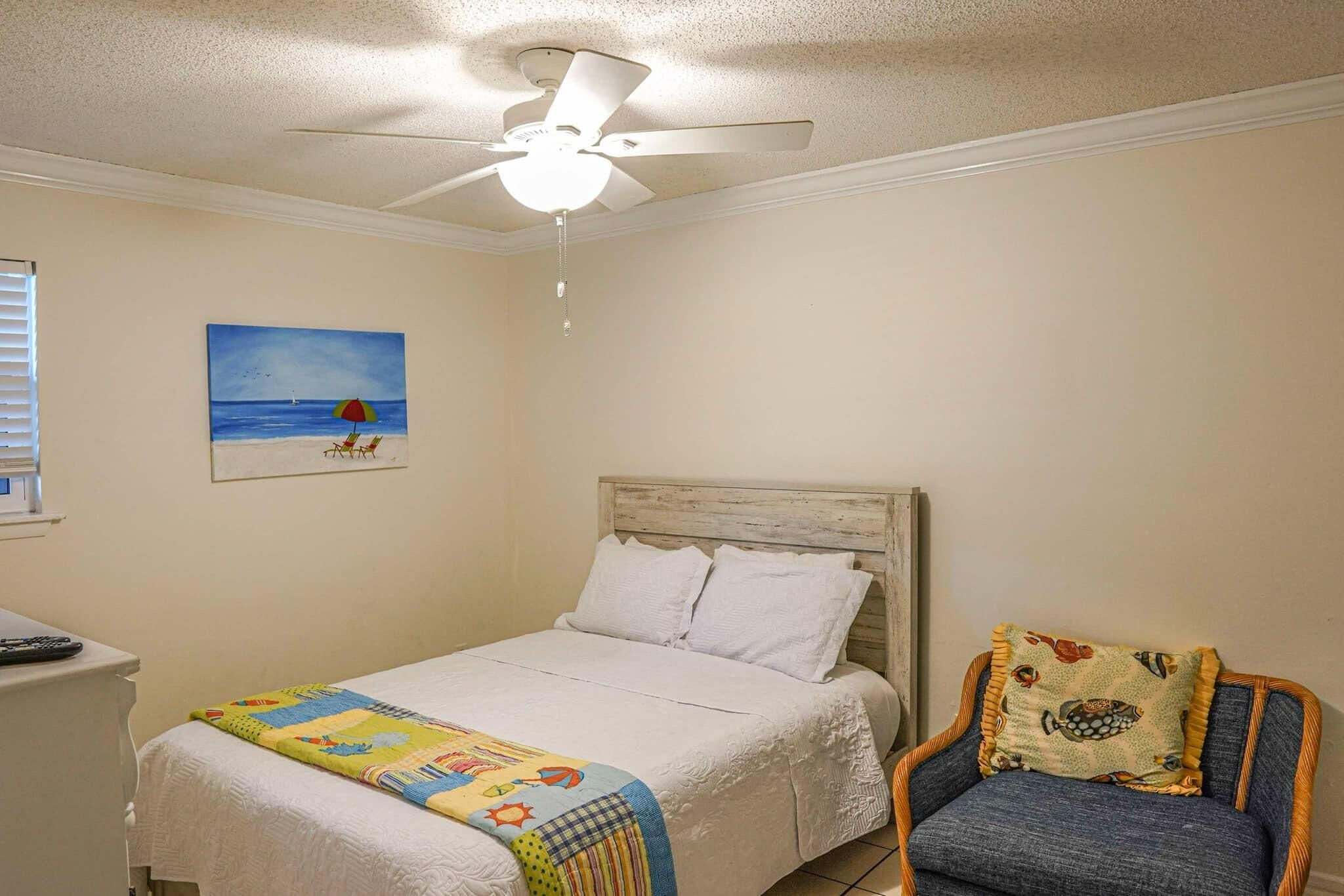 A bedroom with a bed, a chair, a ceiling fan, a painting, and a window.
