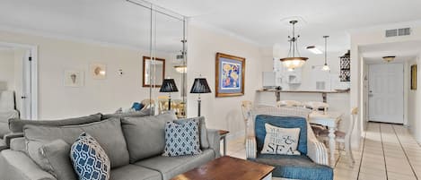 A living room with a sofa, a wooden coffee table, and a wicker chair. There is a kitchen area in the background with a dining table and chairs.