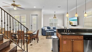 A modern kitchen with a built-in dishwasher, a dining area with a wooden table and chairs, a living room with a sofa and a TV, and a staircase with a ceiling fan. - Sugar Sands (Miramar Beach)