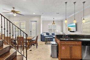 A modern kitchen with a built-in dishwasher, a dining area with a wooden table and chairs, a living room with a sofa and a TV, and a staircase with a ceiling fan. - Sugar Sands (Miramar Beach)