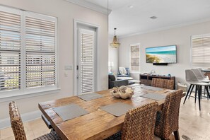 A dining area with a wooden table, woven chairs, and a television displaying a beach scene. - Sugar Sands (Miramar Beach)