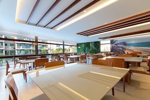 Ein modernes Restaurant mit Holztischen und -stühlen, großen Fenstern und einem Wandbild mit einer Strandansicht.