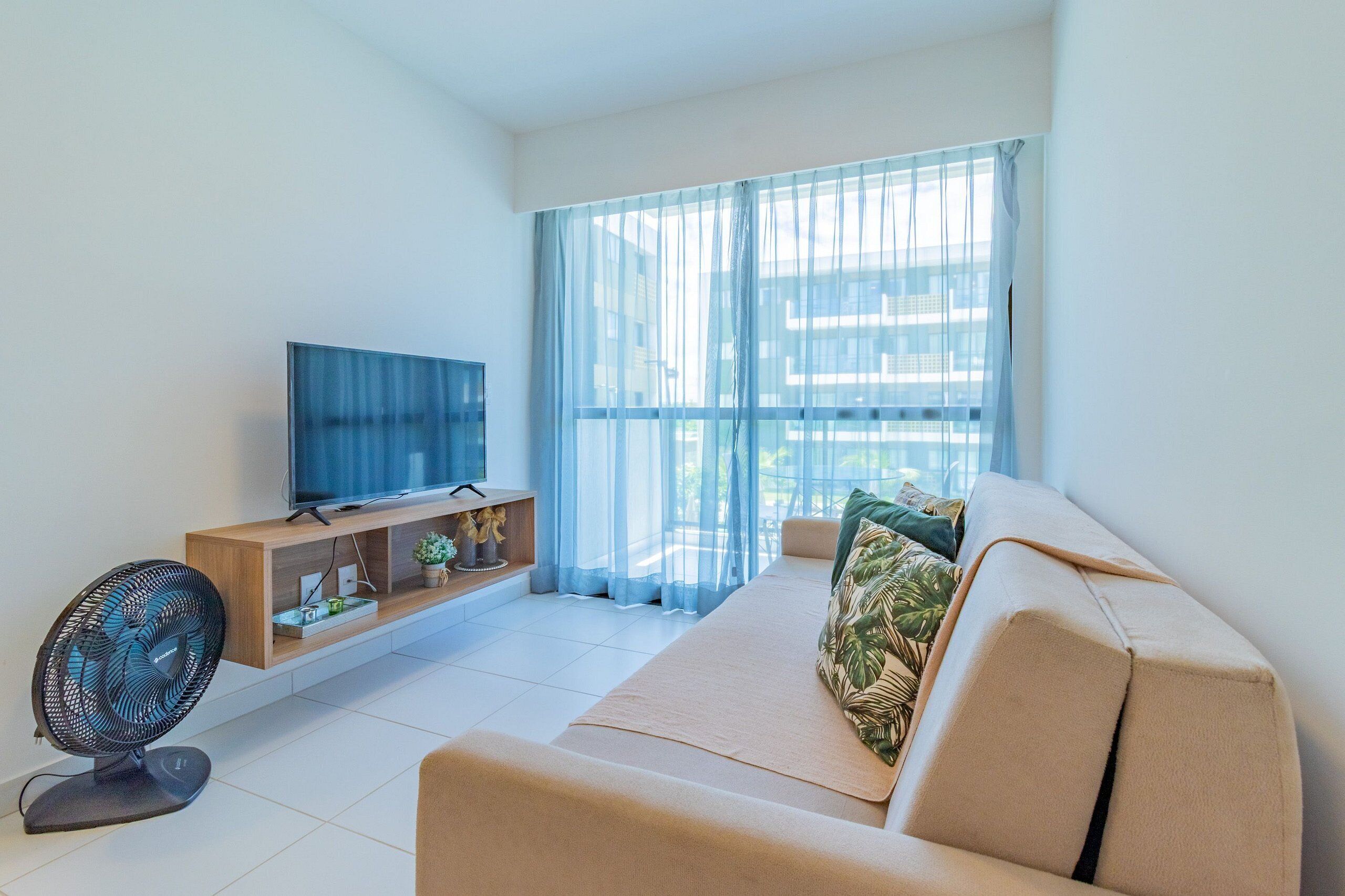 Una sala de estar moderna con un sofá, un televisor, un ventilador y una ventana con cortinas translúcidas.