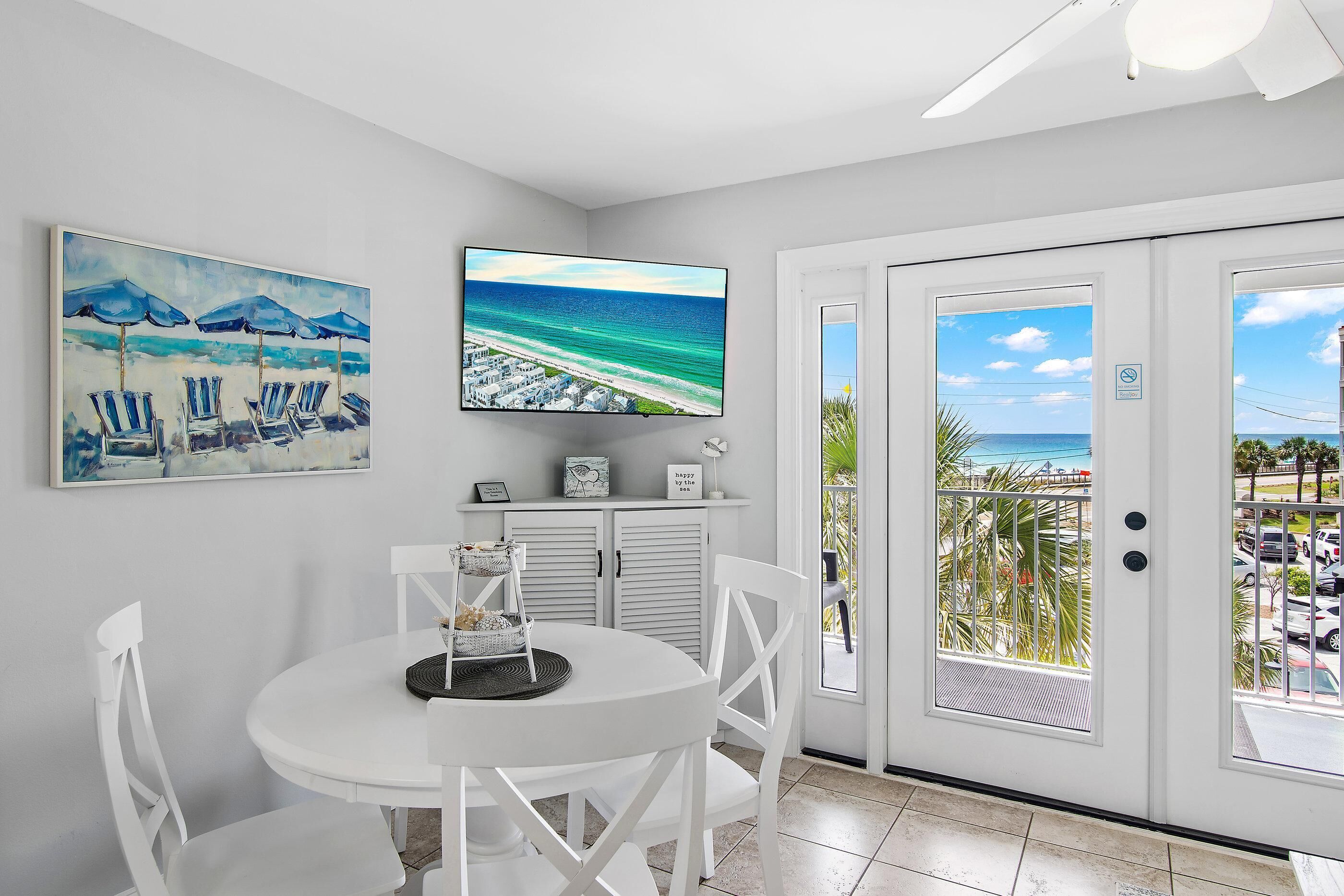 Une pièce avec une table blanche, des chaises, un téléviseur et une porte donnant sur un balcon avec vue sur la plage.