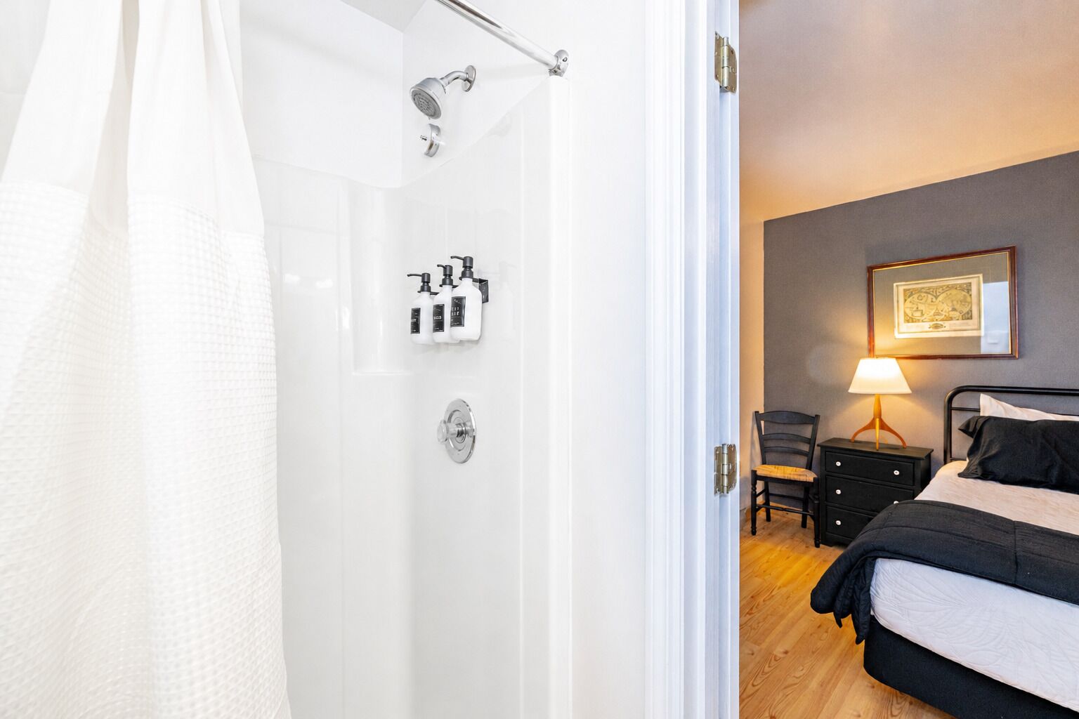 A bathroom with a shower, toilet, and sink, a bed with a bedside table and lamp, and a chair.
