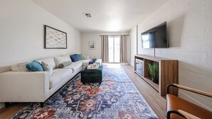 Ein Wohnzimmer mit einer Couch, Hocker, Fernseher und einem gemusterten Teppich.