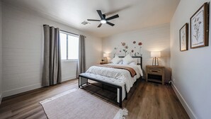 Ein Schlafzimmer mit einem Bett, Nachttischen, einer Bank, einem Deckenventilator und einem Fenster mit Vorhängen.