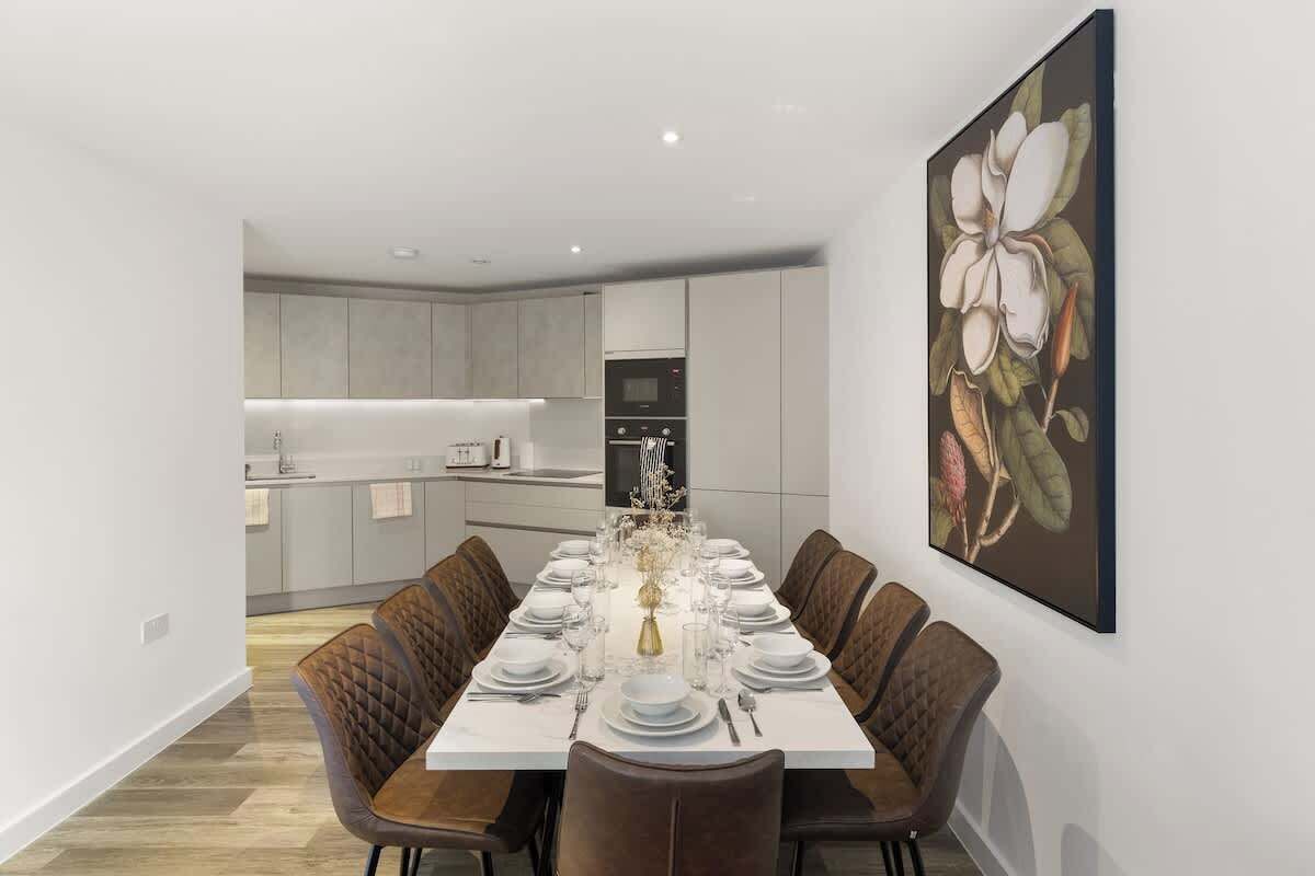 Un comedor moderno con una mesa larga dispuesta para seis comensales, una cocina al fondo y un cuadro floral grande en la pared.