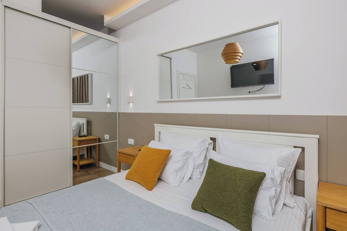 A bedroom with a bed, a bedside table, a nightstand, a mirror, and a wall-mounted television.