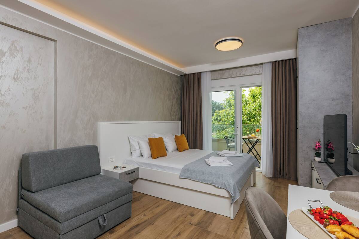 A modern hotel room with a bed, grey sofa, and a dining table with fruit.