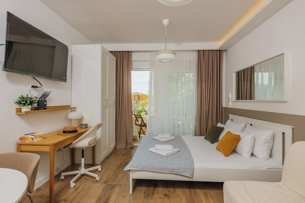 A modern hotel room with a bed, desk, chair, TV, and a view of the outdoors.