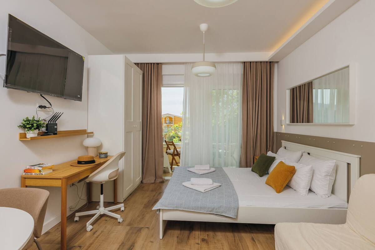 A modern hotel room with a bed, desk, chair, TV, and a view of the outdoors.