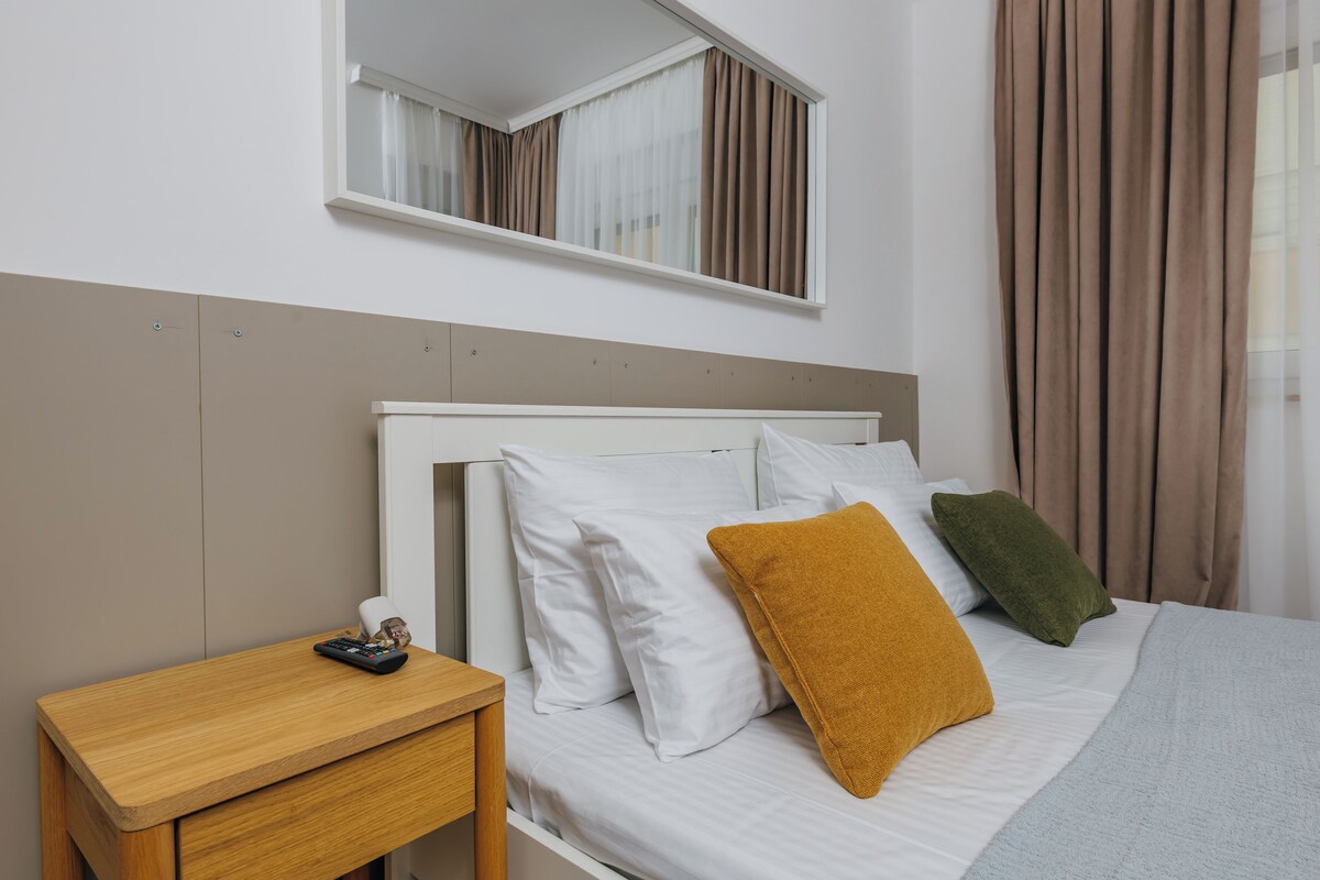 A neatly made bed with white and colored pillows, a wooden nightstand with a remote control, and a window with curtains.