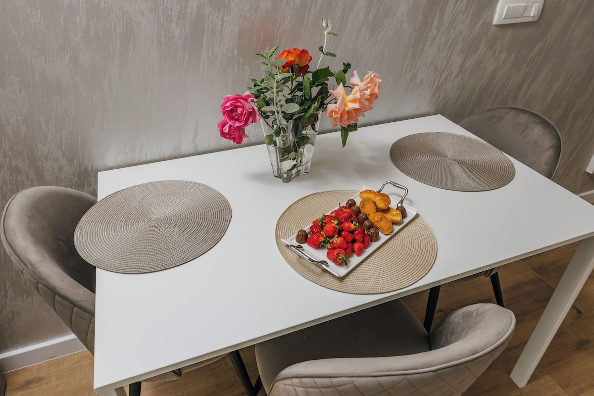Una mesa de comedor con mantel blanco, dos individuales, un florero con flores y un plato de fruta.