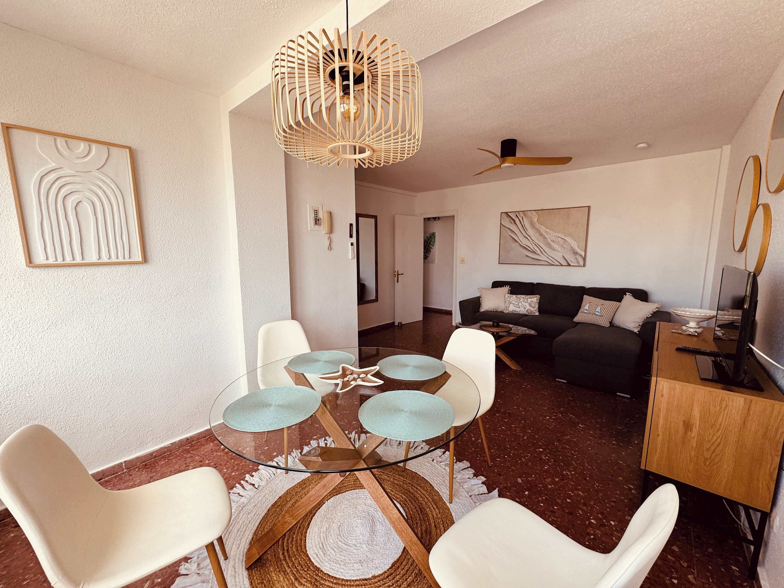 Una sala de estar moderna con una mesa de comedor de vidrio, sillas blancas, un sofá oscuro, un ventilador de techo y obras de arte en las paredes.