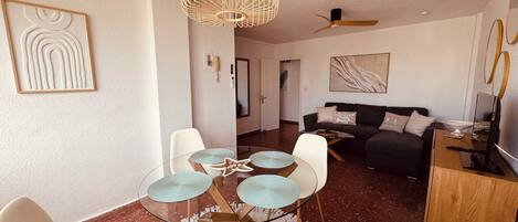 Una sala de estar moderna con una mesa de comedor de vidrio, sillas blancas, un sofá oscuro, un ventilador de techo y obras de arte en las paredes.
