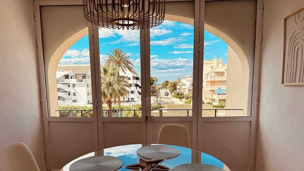 Una habitación con una mesa redonda de vidrio, cuatro sillas, una lámpara en el techo y vistas a una zona residencial a través de amplios ventanales.