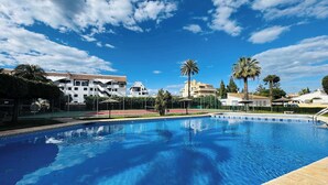 Una piscina bajo un cielo azul despejado, con palmeras y edificios modernos al fondo.