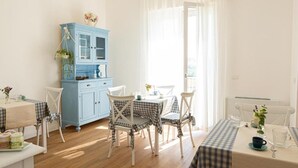 A dining area with a table set for two, a blue cabinet, and a window with curtains. - Dolce Conero Guest House (Camerano)