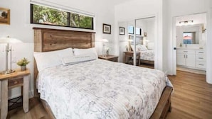 Ein Schlafzimmer mit Holz-Kopfteil, einem Bett mit floralem Überwurf, Nachttischen mit Lampen, einem Fenster mit Blick ins Grüne und einem Badezimmer mit Spiegel und Schränken.