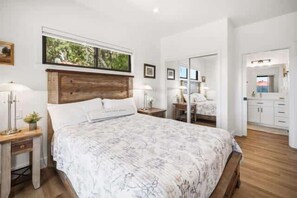 Ein Schlafzimmer mit Holz-Kopfteil, einem Bett mit floralem Überwurf, Nachttischen mit Lampen, einem Fenster mit Blick ins Grüne und einem Badezimmer mit Spiegel und Schränken.