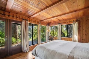 Ein Schlafzimmer einer Holzhütte mit einem großen Bett, mehreren Fenstern und Blick auf den Wald.