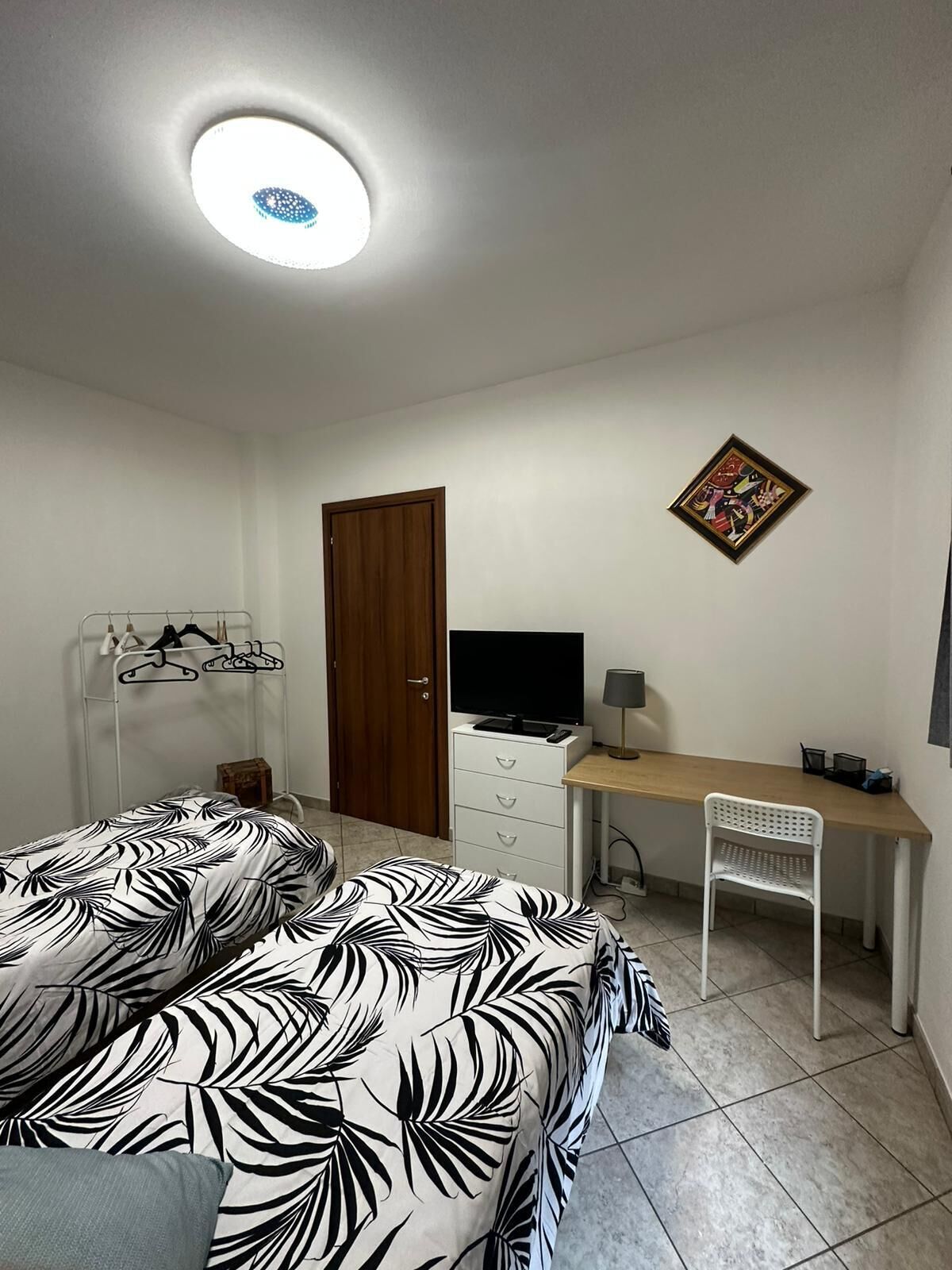 A bedroom with a bed, a desk, a chair, a TV, a door, and a ceiling light.