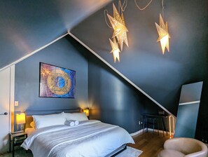 Ein Schlafzimmer mit schrägen Wänden, einem Bett mit weißer Bettwäsche, einem Wandbild und zwei wandmontierten Sternlampen.