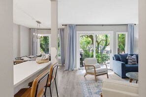 Ein modernes Wohnzimmer mit Essbereich, großen Fenstern mit Vorhängen, einer Couch und einem Sessel.