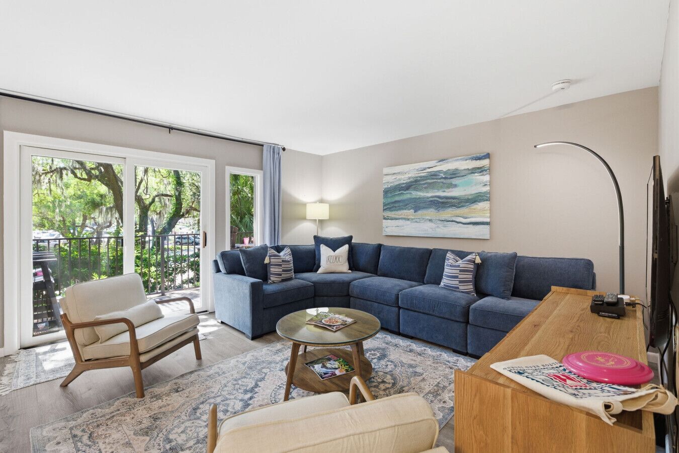 Ein Wohnzimmer mit blauem Sofa, einem Holztisch, einem beige farbenen Sessel, einem Glastisch und einem Fernseher.