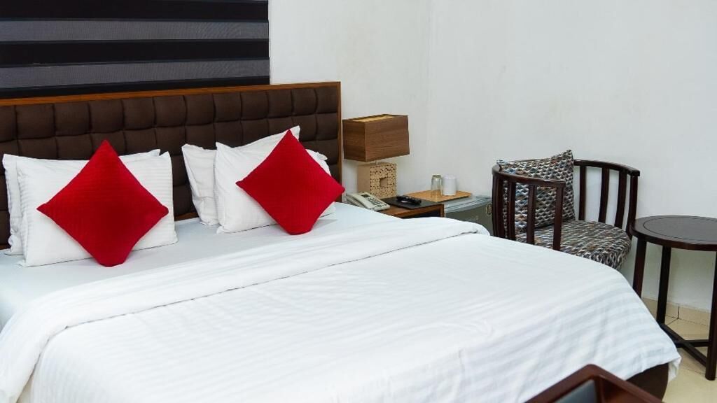 A hotel room with a large bed, two red pillows, a wooden headboard, a bedside table with a lamp, a chair, and a small round table.