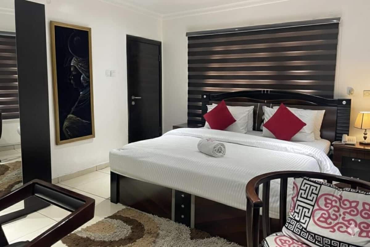 Un dormitorio ordenado con una cama grande, una mesita de noche, una silla y un cuadro en la pared.
