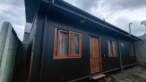 A small, dark-colored building with two windows and a door, a wooden bench outside, and a cloudy sky.