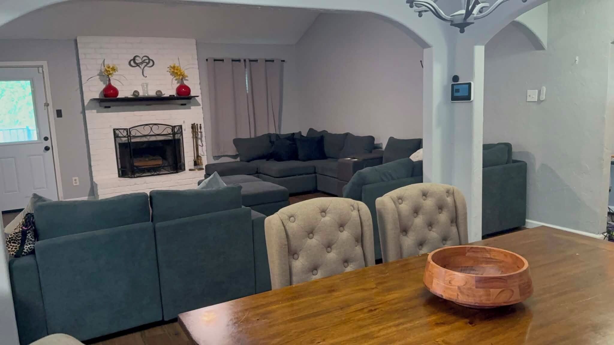 Una sala de estar con chimenea, sofás grises, una mesa de comedor y un cuenco de madera, y una lámpara de araña moderna.