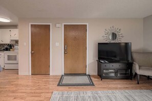 A living room with a TV, a sofa, a wooden door, and a kitchen in the background.