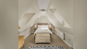 Une chambre avec un plafond en pente, un lit recouvert de draps blancs, deux lampes de chevet, une commode en bois et un tapis à motifs.