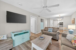 A living room with a flat-screen TV, a ceiling fan, a light blue cabinet, a brown sofa, a coffee table, and a dining area in the background. - Beach Access, Pool & Tennis - Sleeps 6 (Hilton Head Island)