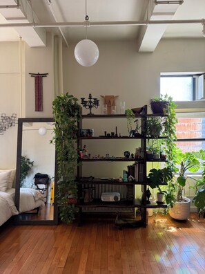 A room with a wooden floor, a bookshelf with plants, a hanging light fixture, and a window with a view. - Studio Loft in the heart of NYC  - 5 min walk to time square (New York)