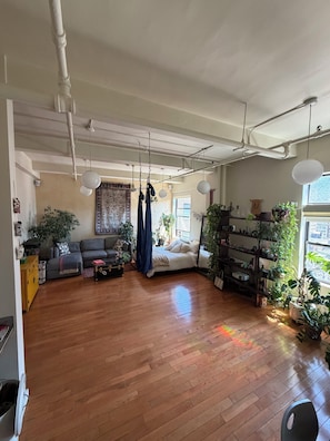 A spacious living room with a wooden floor, a sofa, a bed, hanging plants, and a window with a city view. - Studio Loft in the heart of NYC  - 5 min walk to time square (New York)