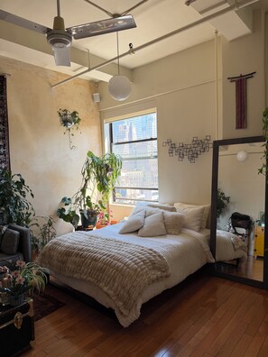 A bedroom with a bed, a ceiling fan, a window with a city view, and several potted plants. - Studio Loft in the heart of NYC  - 5 min walk to time square (New York)