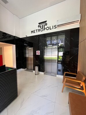 Un área de recepción moderna con un letrero que dice 'METROPOLIS', un bote de basura, un ascensor y una zona de estar con sillas.